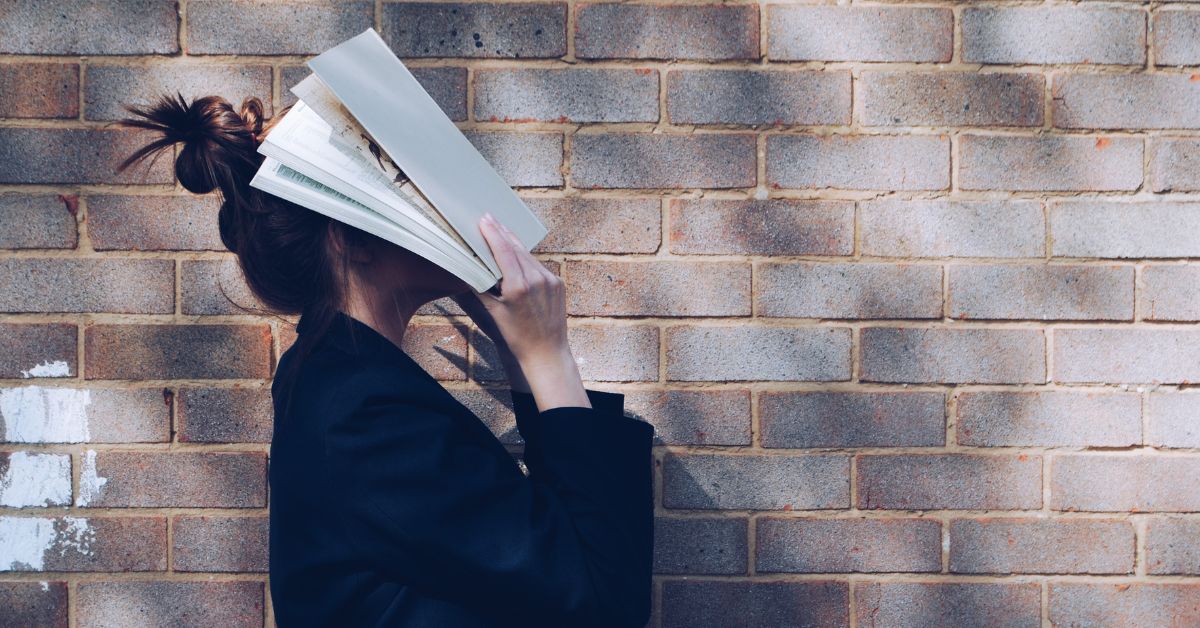 School Girl with Book in front of natural rustic red brick background holding book up to her face.